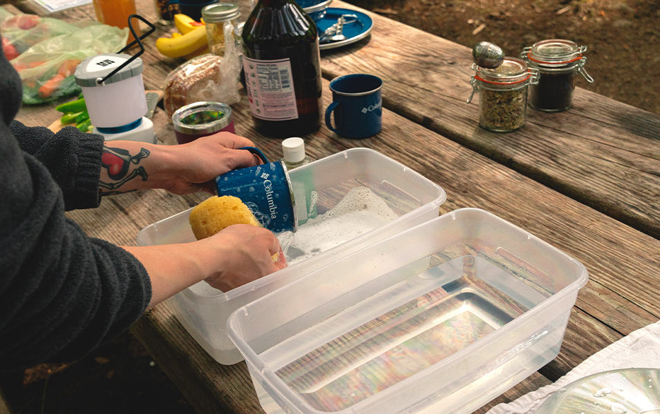 A close-up shot on a pair of hands washing a cup in two plastic bins that are set up on a camping picnic table.