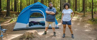 A man and a woman stand in the woods facing the camera with a blue tent and trees in the background.