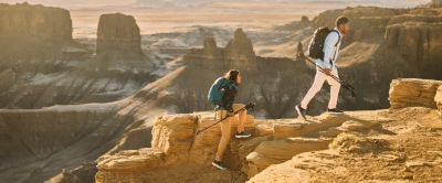 Two hikers in sun-protective gear hike above a vast desert canyon.
