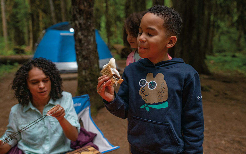 A boy in a navy blue sweatshirt eats S’mores in the woods as his mom skewers a marshmallow in the background. 
