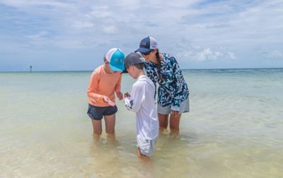 A boy and a girl wade in the water holding a tiny fish as a woman peers into their hands looking at it and smiling.