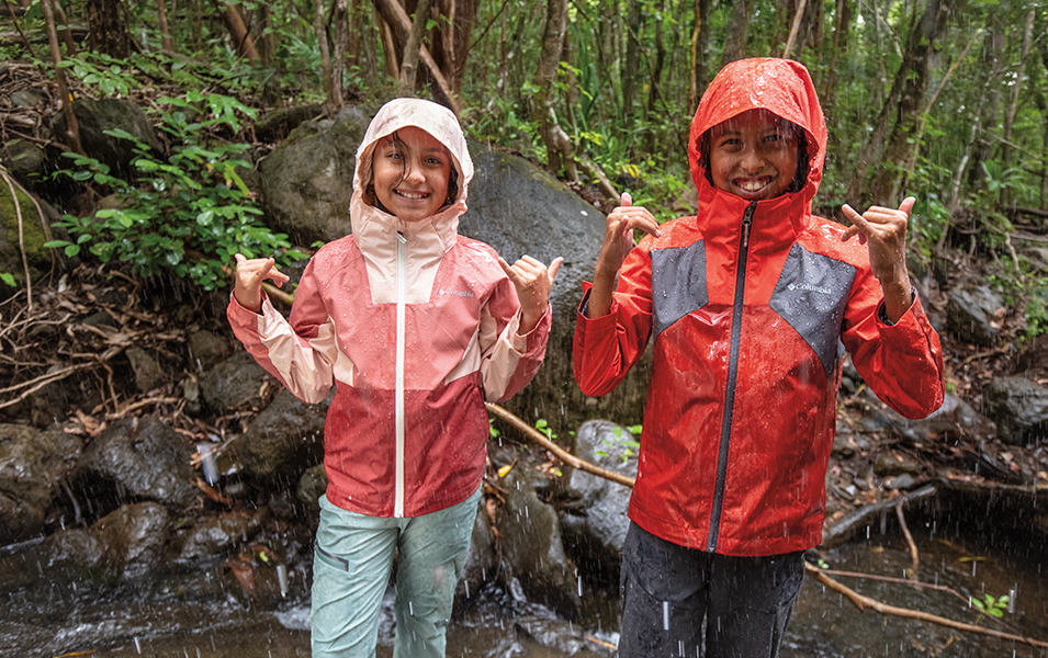 Two girls wearing Columbia Sportswear jackets stand in the rain smiling in the middle of the forest. 