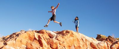 A pair of kids jump across rocks on top of a scenic canyon on a sunny day.