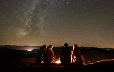 Four people are sitting on a fallen log, gazing into a glowing campfire. Behind them, stars light up the night sky.