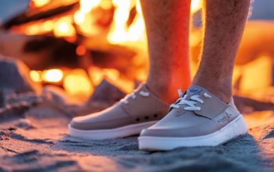 A man wearing boat shoes and no socks stands in front of a flickering campfire.