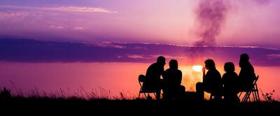 Six people sit in chairs around a campfire as a brilliant pink and purple sunset reflects in the lake behind them.