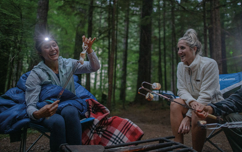 Two women sit around a campfire roasting marshmallows and smiling.