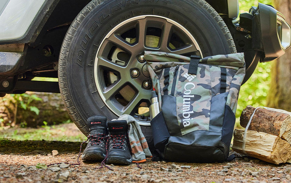A Columbia Sportswear drybag sits next to a vehicle, alongside a pair of hiking boots and campfire wood