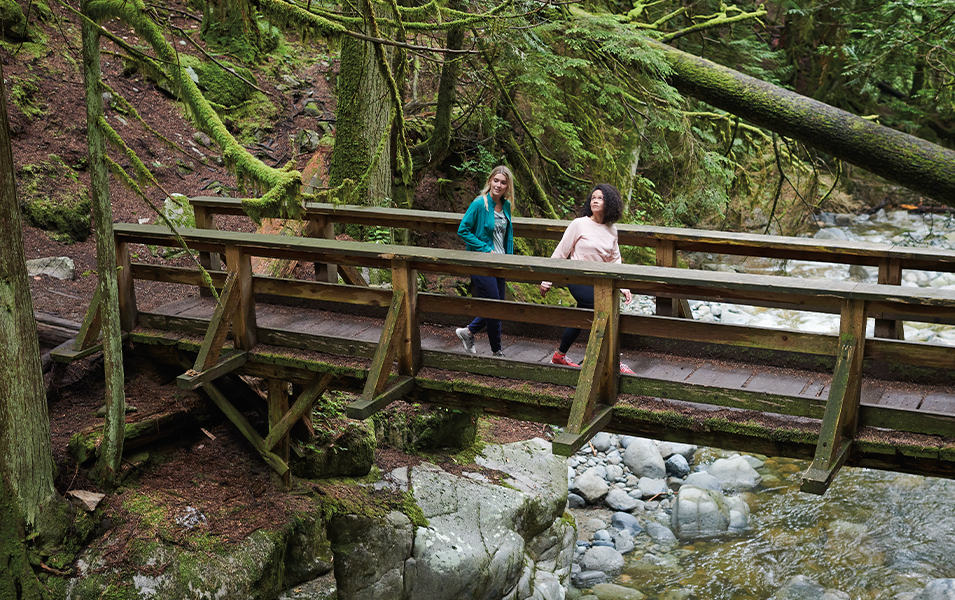 Two women hike across a wooden bridge in a mossy forest with a rushing creek beneath them.