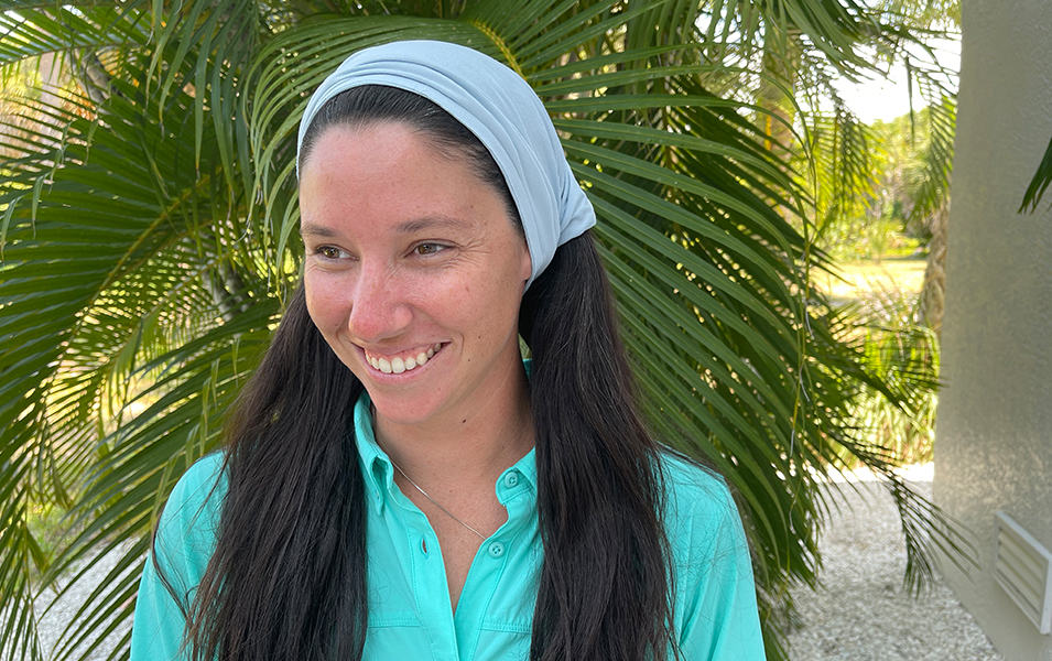 Woman wearing neck gaiter like a headscarf next to a palm tree. 