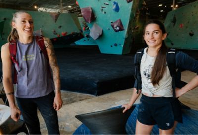 Alex Johnson and Kyra Condie smile for the camera inside the climbing gym, both with backpacks on, ready to go.