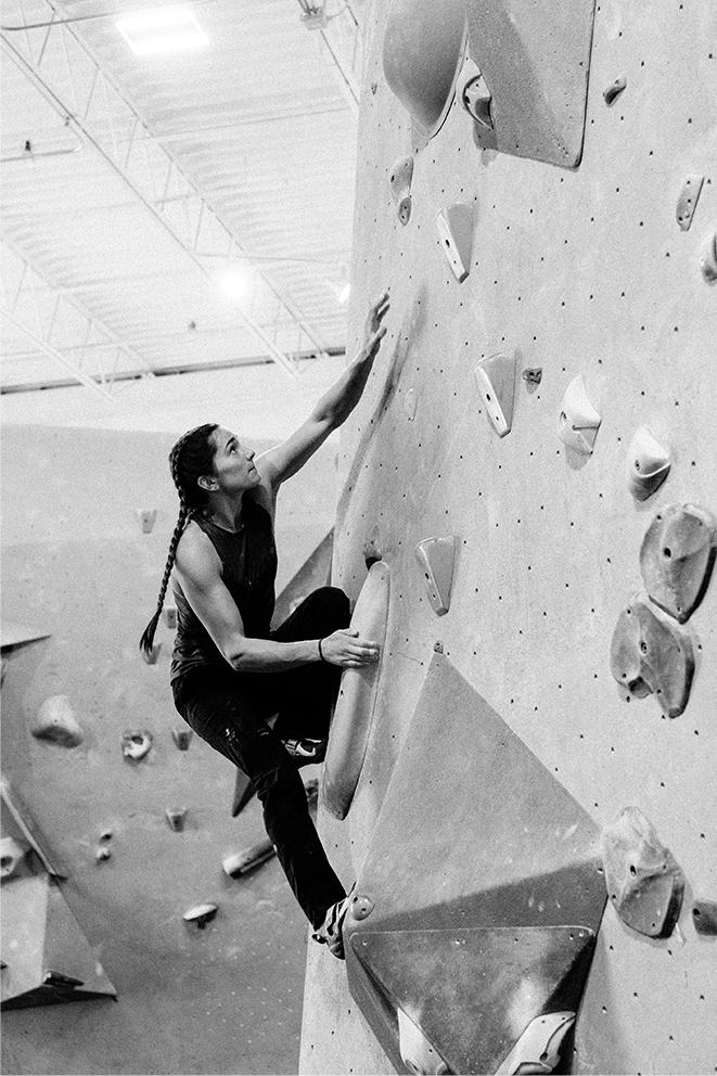Black and white image of Kyra Condie bouldering, reaching up for her next move.