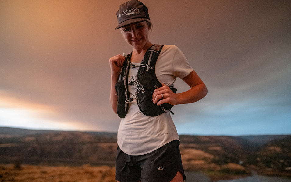 A trail runner in a white shirt and black hydration vest stops to adjust her gear while facing the camera.