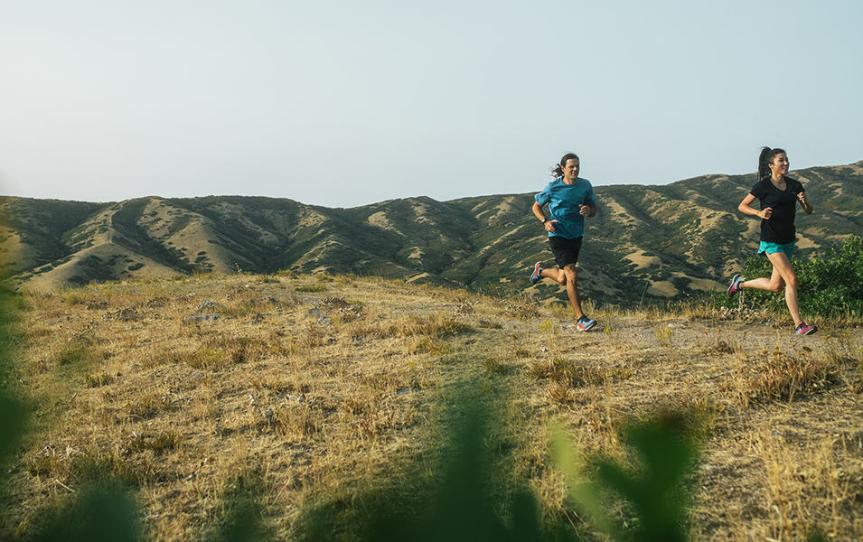 A man and a woman run along an open trail in the sun with grassy plains in the background.