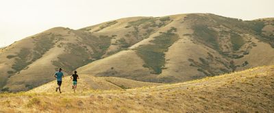 Two trail runners making their way across a trail on a sunny day in the mountains. 
