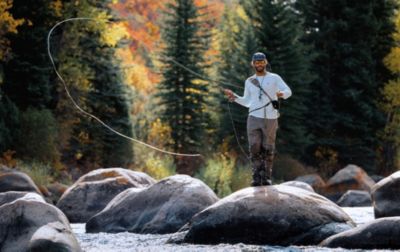 An angler with a fly rod stands on a boulder, casting his line into a mountain creek. 
