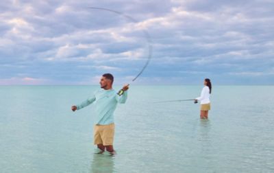 Standing knee-deep in the shoals, two saltwater anglers wear sun-protective hoodies and quick-drying shorts as they cast their lines. 
