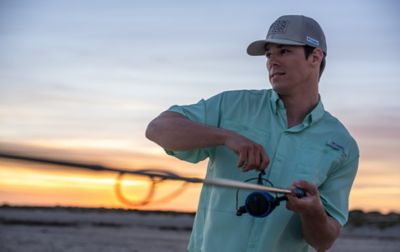 As the sun sets, a man with a reel slowly takes in his fishing line. 