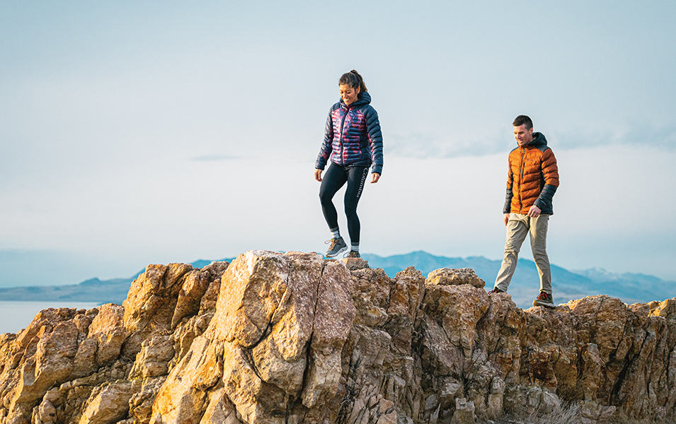 A woman in a blue-and-purple puffer jacket stands in front of a man wearing an orange hiking jacket as the two walk across a rocky cliff with mountains in the background. 