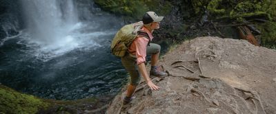 A woman wearing a pink hiking shirt and pale green pants climbs up a rock in a scenic forest with a waterfall in the background. 