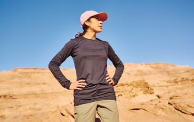 A woman wearing a brimmed ball cap and a long-sleeve sun-protective shirt stands with her hands on her hips. 