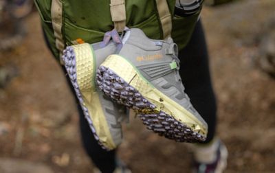 A pair of Columbia Sportswear hiking boots hang from a hiker’s backpack as they walk on a trail 
