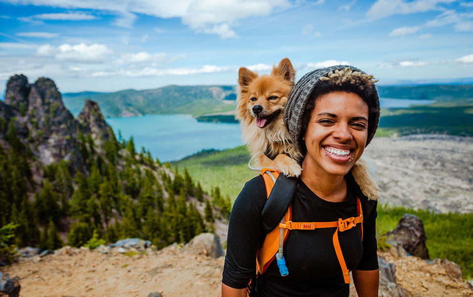 A woman stands atop a beautiful hiking trail with trees and mountains in the background, and her small dog perched on her shoulder.  