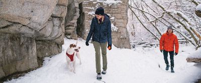  A pair of hikers treks through a snowy landscape with their pup at their side. 
