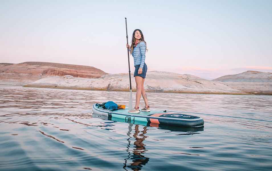 A woman wearing a blue-and-white Columbia Sportswear shirt stands on a paddleboard in a scenic river. 