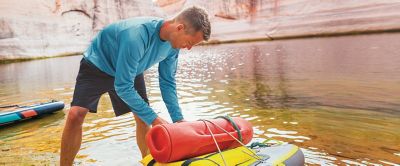 A man wearing a blue Columbia Sportswear shirt attaches gear to a standup paddleboard with a scenic river in the background. 