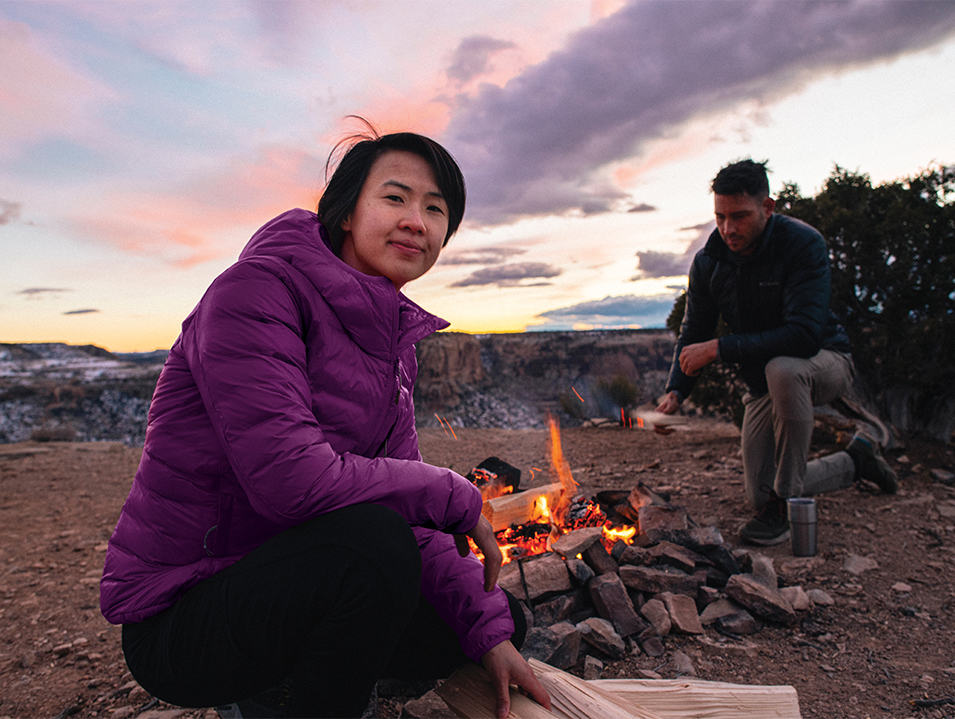 A woman smiles at the camera while tending in front of campfire as a man crouches on a knee in the background.