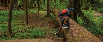 A boy wearing a red Columbia Sportswear backpack climbs over a log in a scenic forest.