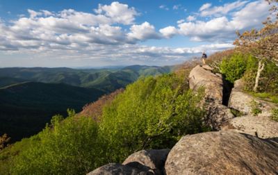 A hiker stands on a large boulder, gazing at rolling ridges and valleys in the distance.