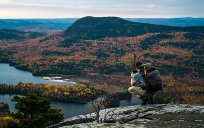 A hiker with a big backpack and hiking poles crouches on a rocky ridge, photographing a lake and tree-covered knoll.