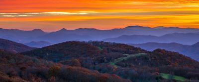 Rolling hills, autumn foliage, and patches of green meadow are framed by a spectacular orange sunset.