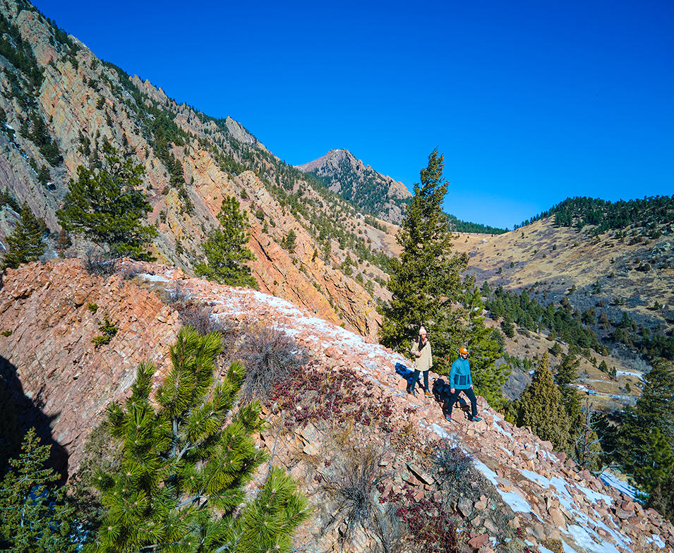 A pair of hikers walk down a steep, rocky trail in a beautiful mountainous setting. 