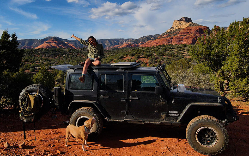 A woman on an overlanding adventure sits on top of a Jeep in the high desert smiling with her arm in the air as her dog looks up at her. 