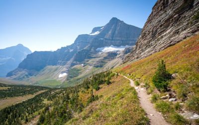 A dirt footpath traverses an alpine slope. The trail leads into a wide valley surrounded by steep mountains with several snowfields high up on their flanks.