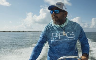 A bearded man wearing a ball cap and sunglass is driving a boat.