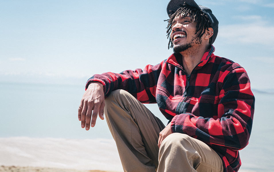  A man sits on a beach wearing a flannel shirt with a red-and-black buffalo check pattern. 