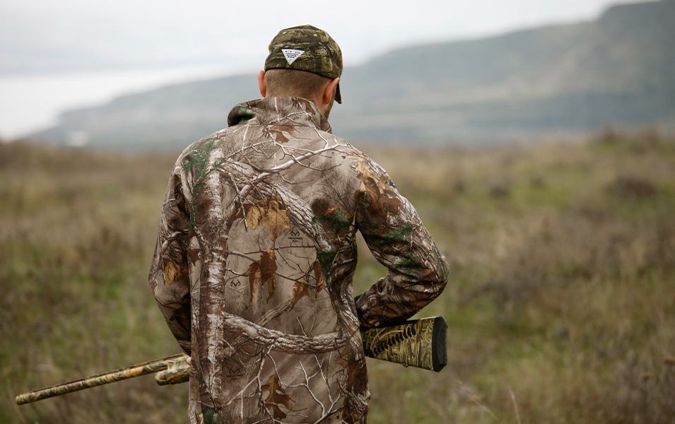 A hunter stands in a field holding a rifle with his back to the camera.