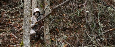 A hunter crouches behind a tree wearing full-body camouflage in the woods. 
