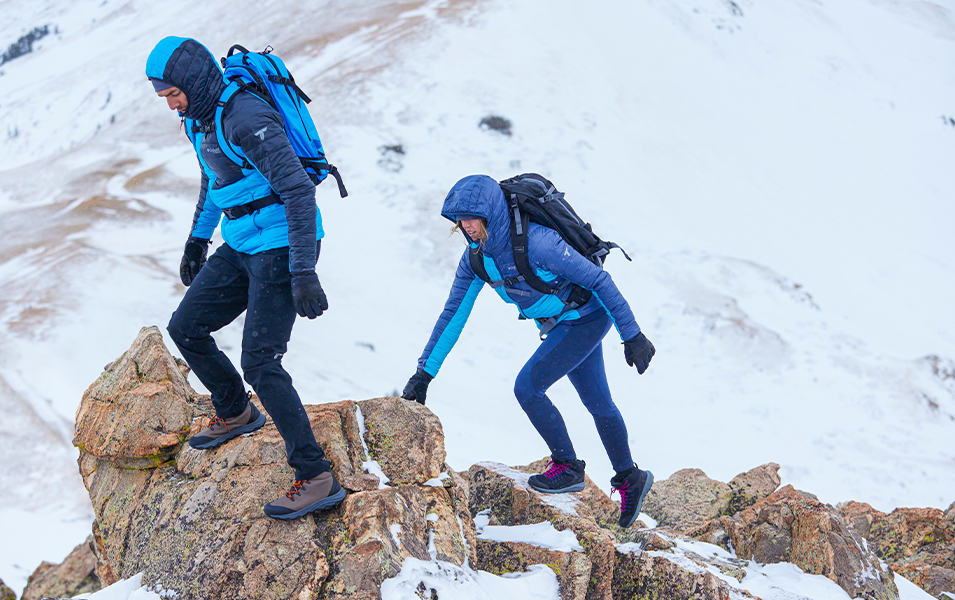A man and woman hike across jagged snow-covered rocks wearing insulated puffer jackets with backpacks on.