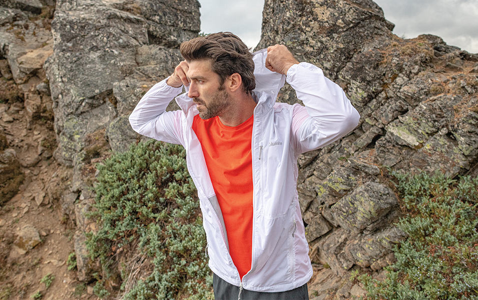 A man fixes the hood of his windbreaker jacket while standing in front of a jagged rock outcrop.