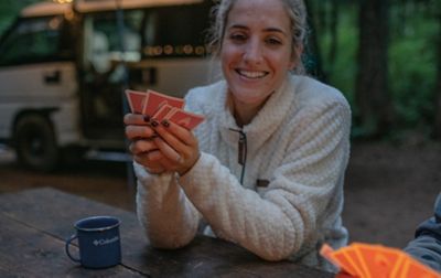 A woman sits at a campsite picnic table playing cards wearing a cozy fleece jacket.