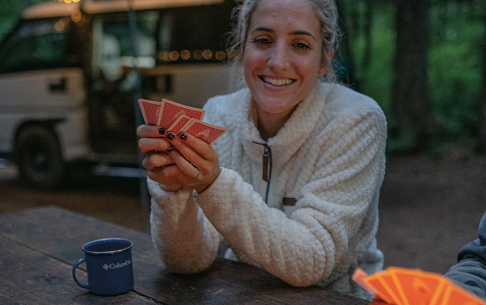 A woman sits at a campsite picnic table playing cards wearing a cozy fleece jacket.
