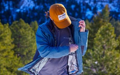 A man stands in snow dusted forest zipping up the inner liner of his Interchange jacket.