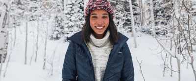 A woman is standing in the foreground of a snow-covered forest wearing a puffer jacket.