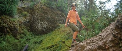 A hiker stands on a rocky trail in a lush green forest looking down at the view behind her. 