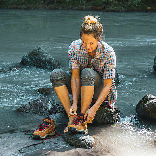 A woman putting hiking boots on next to a river.